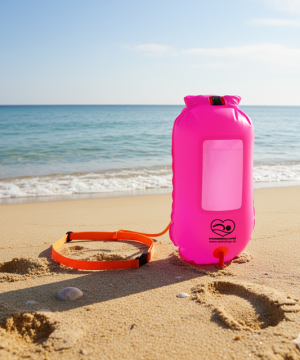 En lyserød OpenWater Swimming Buoy - Havtaske - 20L (Pink) med en orange strop ligger på sandstranden ved det rolige blå hav, hvilket gør den til den perfekte pålidelige tørpose og bøje til svømmeeventyr i åbent vand.
