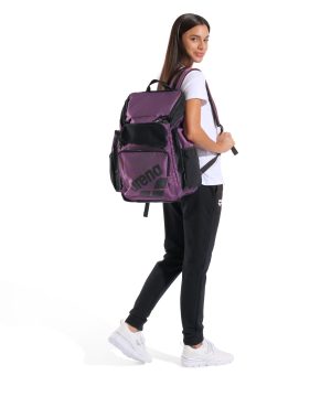 En kvinde står og kigger tilbage, klædt i en hvid t-shirt, sorte bukser og hvide sneakers, og hun bærer den store lilla Arena One Go Backpack 45L AO (Scratched) med sorte detaljer.