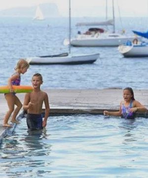 Tre børn hygger sig ved vandet: En pige står ved bassinkanten med en Zoggs Badering (2-3 år), mens en anden pige og en dreng smiler i bassinet. To sejlbåde ligger for anker i baggrunden.