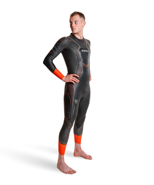 En person står barfodet på en hvid baggrund iført Zone3 Vanquish X Våddragt - Herre våddragt med orange detaljer på håndled og underben, hænderne på hofterne og kigger til siden.