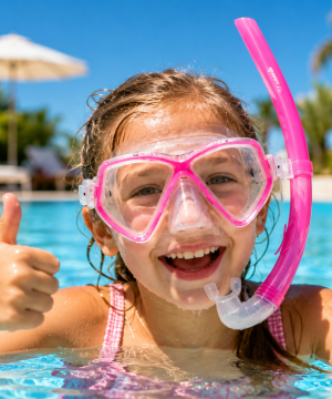 Et smilende barn iført Mares Zephir Combo Snorkelsæt (Pink) – Junior giver tommelfingeren op i en svømmehal, med palmer og en parasol i baggrunden på en solrig dag.